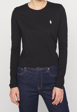 Persona con una camiseta ajustada de manga larga negra con un pequeño logo blanco de un jugador de polo en el pecho y jeans azul oscuro.