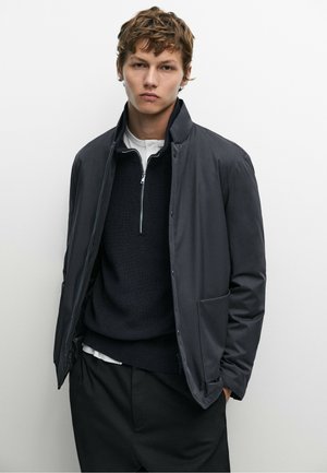 Giovane uomo con capelli corti mossi che indossa un maglione nero con zip sotto una giacca scura e pantaloni neri, in piedi davanti a uno sfondo bianco uniforme.