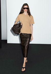 T-shirt beige, gonna in pelle marrone e borsa nera. La modella indossa occhiali da sole oversize e ciabatte nere, posando su uno sfondo semplice.