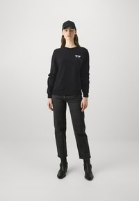 Svart hettegenser med lange ermer og rund hals, med et lite hvitt grafisk motiv på brystet, kombinert med mørkejeans og svarte ankelstøvler.