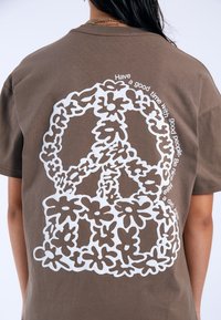 Camiseta de algodón marrón con un gran símbolo de paz blanco rodeado de contornos florales y texto que envuelve el diseño en la parte posterior.