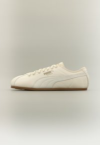 Χαμηλού προφίλ κρεμ αθλητικό παπούτσι Puma Tackle με σουέντ ρίγα και καφέ σόλα, εμφανισμένο από την πλευρά σε ουδέτερο βαθμωτό φόντο.