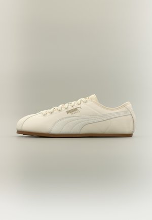 Sneaker Puma Tackle color crema a profilo basso con striscia in camoscio e suola marrone, mostrata di lato su uno sfondo sfumato neutro.