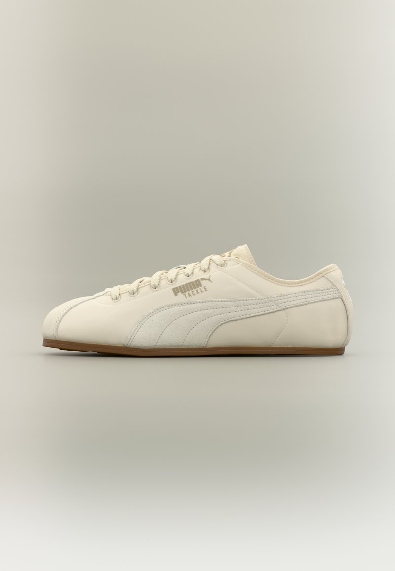 Χαμηλού προφίλ κρεμ αθλητικό παπούτσι Puma Tackle με σουέντ ρίγα και καφέ σόλα, εμφανισμένο από την πλευρά σε ουδέτερο βαθμωτό φόντο.