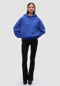 Felpa blu oversize in materiale felpato con tasca frontale e logo ricamato. Abbinata a pantaloni neri attillati e décolleté a punta.