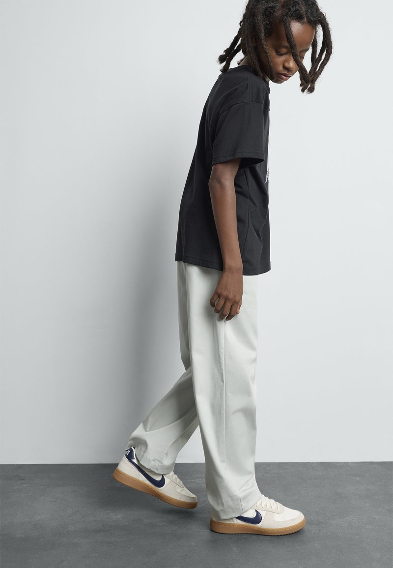 Persona con rastas, vistiendo una camiseta negra, pantalones gris claro y zapatillas blancas con un logo azul, apoyado contra una pared gris lisa.