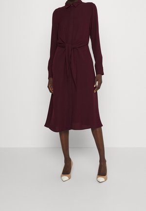 Kobieta w bordowej sukience z długim rękawem, sięgającej do kolan, z wiązaniem z przodu oraz beżowych szpilkach z białymi końcówkami.