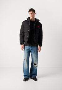 Giacca puffer nera con cappuccio e toppa rossa, abbinata a jeans larghi sbiaditi di colore blu chiaro. Tessuto morbido e sezioni trapuntate.