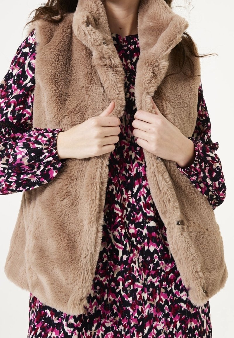 Kobieta ubrana w beżowy kamizelkę z sztucznego futra, na którą ma założoną długą sukienkę w kwiaty w kolorach purpurowym, czarnym i białym, poprawiająca kołnierz kamizelki.