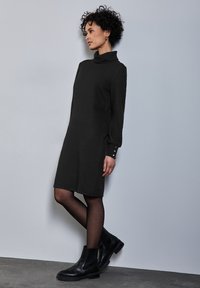 Černé rolákové šaty s dlouhými rukávy a knoflíkovými detaily na manžetách, které jsou doplněny černými kotníkovými botami a nošeny přes průhledné černé punčochy.
