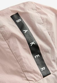 Ljust rosa tyg med en slät textur som har en svart rektangulär lappar som visar ordet "BAKER" i vita versaler.