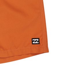 Pantaloncini arancioni realizzati in tessuto leggero, con orlo piatto e un patch logo in bianco e nero sul lato.