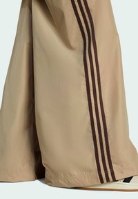 Béžové teplákové nohavice s tromi hnedými pruhmi po stranách, vyrobené z ľahkej látky, ktoré majú voľný strih a lem s elastickým detailom.