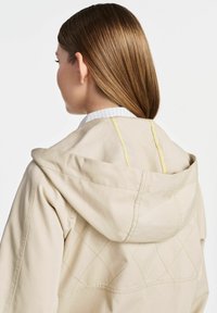 Beżowa kurtka z kapturem z żółtymi przeszyciami, z diamentowym wzorem na plecach. Gładki materiał i casualowy krój.