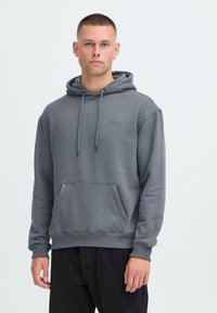 Hombre con cabello corto que lleva una sudadera gris con bolsillo frontal y pantalones negros, de pie frente a un fondo claro y liso.