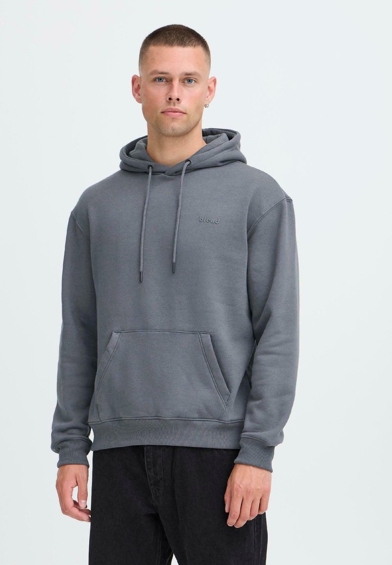 Hombre con cabello corto que lleva una sudadera gris con bolsillo frontal y pantalones negros, de pie frente a un fondo claro y liso.