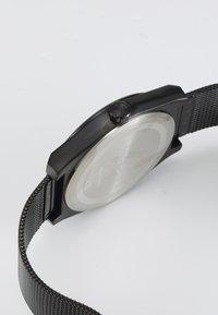 Reloj con correa de malla negra, caja redonda, parte trasera de metal texturizado y corona negra. Presenta un diseño minimalista y una resistencia al agua de 5 bares.