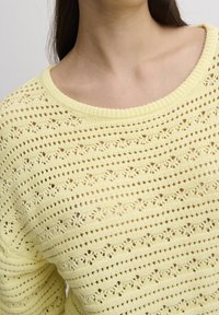Suéter amarillo de punto que presenta un diseño de patrones calados con rayas horizontales. El escote es amplio y acanalado, con un acabado texturizado.