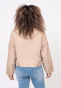Veste beige courte avec des manches larges, texture lisse et détails contrastants aux épaules. Associée à un jean en denim bleu clair.