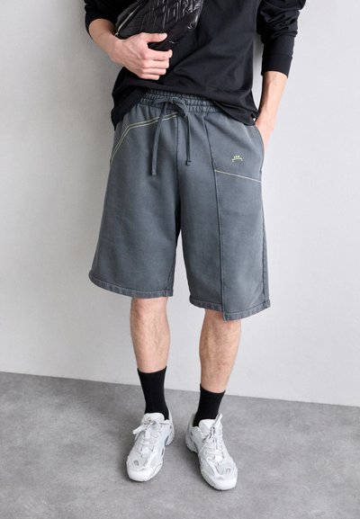 Graue Sportshorts mit einem Kordelzugbund, versehen mit neon-gelber Naht und Seitentaschen. Getragen mit schwarzen Socken und grauen Turnschuhen.