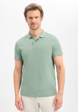 Mann trägt hellgrünes, strukturiertes kurzärmeliges Poloshirt und beige Hose, steht vor einem schlichten weißen Hintergrund.