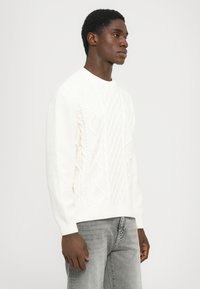 Vit stickad tröja med ett strukturerat diamantmönster, rund halsringning och långa ärmar. Bärs med grå jeans.