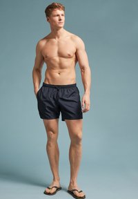 Marineblauwe zwemshorts van lichtgewicht stof met een elastische tailleband en twee zijzakken, gecombineerd met zwarte teenslippers.