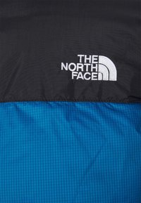 Czarna i niebieska tkanina izolacyjna z pikowanym wzorem w niebieskiej części. Białe haftowane logo "THE NORTH FACE" wyeksponowane w widocznym miejscu.