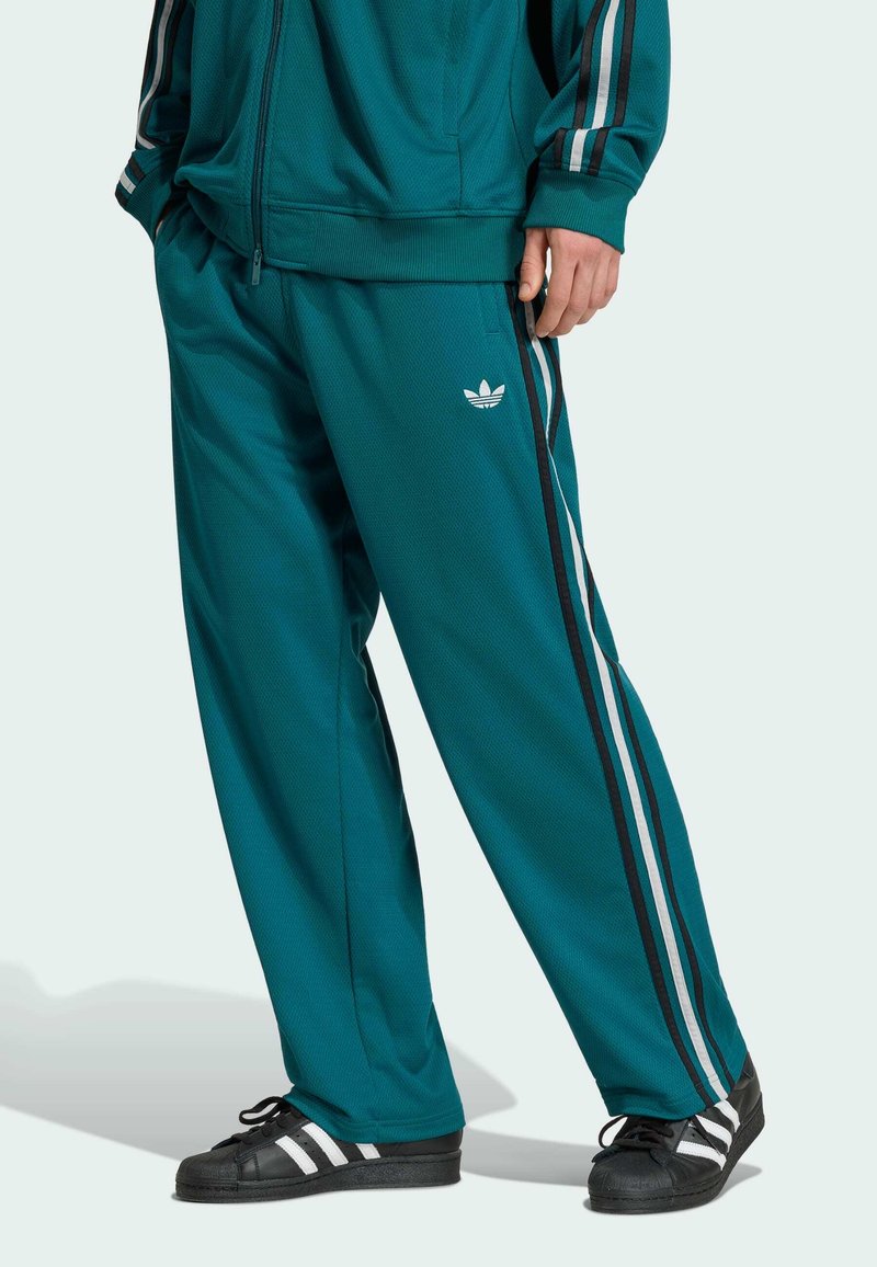 Türkisfarbene Adidas-Trainingshose und passende Jacke mit weißen und schwarzen Seitenstreifen, kombiniert mit schwarzen Adidas-Sneakern mit weißen Streifen.