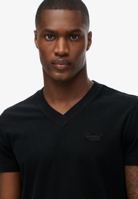 Sort bomulds t-shirt med v-hals design, der har et syet logo på venstre bryst. Stoffet er glat med en tætsiddende silhuet.