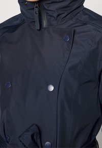 Veste imperméable marine avec un col montant, un design croisé, une texture lisse et de grands boutons brillants pour la fermeture.