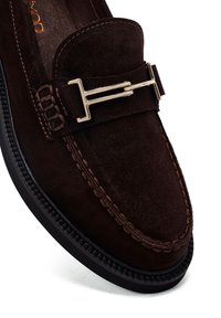 Los mocasines de ante marrón presentan una hebilla de metal dorado, costuras en contraste y una suela de goma texturizada para mayor agarre. Parte superior suave con diseño sin cordones.