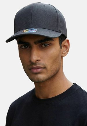 Homme à la barbe taillée et aux yeux foncés, portant une casquette de baseball unie et sombre ainsi qu'un t-shirt noir à col rond, regardant droit devant lui.