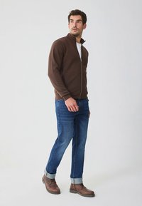 Veste marron zippée, chemise blanche en dessous, jean bleu avec ourlets retroussés, bottines marron. Tissu lisse, coupe décontractée, design casual.