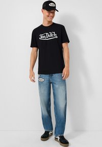 Black cotton t-shirt with white “Von Dutch” logo. Light blue straight-leg jeans with logo patches. Black cap and black sneakers with white accents.