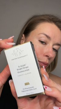Frau hält die Verpackung des Hautpflegeprodukts Drunk Elephant B-Goldi Bright Drops, zeigt Text und Logo, trägt weiße Handschuhe und Herz-Nageldesign.