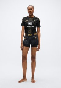 Črna UFC športna oblačila z oprijetim kratkim rokavom, okrašenim z zlatimi poudarki, in ujemajočimi se kratkimi hlačami, oba z grafičnimi vzorci in logotipom.