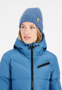 Blaue Daunenjacke mit hohem Kragen, Reißverschlusstaschen und schwarzen Akzenten; kombiniert mit einer strukturierten blauen Strickmütze mit einem Etikett.