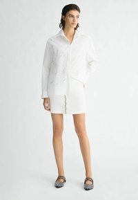 Camicia bianca con dettagli a traforato, abbinata a una mini gonna bianca con un pannello frontale decorativo a bottoni e ballerine nere con decorazioni.