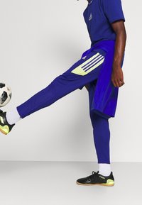 Osoba w niebieskim stroju sportowym Adidas kopie czarno-białą piłkę nożną w pomieszczeniu, nosząc czarne i neonowo-żółte buty do futbolu halowego.