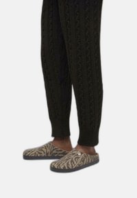 Zapatillas de lana sin cordones con un patrón de cebra marrón y beige, combinadas con pantalones de punto oscuros que presentan texturas de trenzas prominentes.