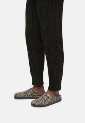 Uld sliders med et brunt og beige zebra-mønster, kombineret med mørke strikkede bukser med markante kabelstrukturer.