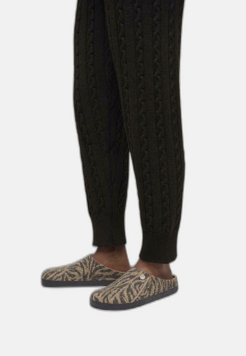 Zapatillas de lana sin cordones con un patrón de cebra marrón y beige, combinadas con pantalones de punto oscuros que presentan texturas de trenzas prominentes.