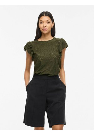 Femme portant un chemisier en dentelle ajourée vert olive à manches courtes et un short noir taille haute, longueur genou, sur mesure, avec les mains dans les poches.