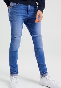 Jeans ajustados de mezclilla azul con diseño clásico de cinco bolsillos, costuras en contraste y dobladillos acampanados, combinados con zapatillas blancas.