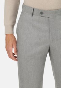 Sivé nohavice v herringbone vzore s úzkym strihom, majú opasok, zapínanie na gombík a hladkú textúru látky. Ruka položená na stehne.