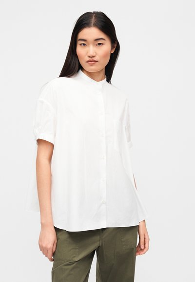 Λευκό πουκάμισο με κουμπιά και όρθιο γιακά, κοντά μανίκια, κατασκευασμένο από ελαφρύ ύφασμα. Διαθέτει τσεπάκι στο στήθος και φαρδιά εφαρμογή.