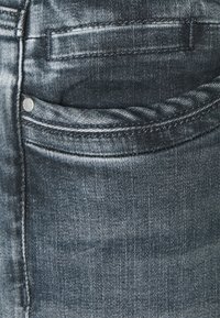 Gros plan sur la poche d'un jean en denim bleu foncé avec des coutures apparentes, un rivet et la texture du tissu.