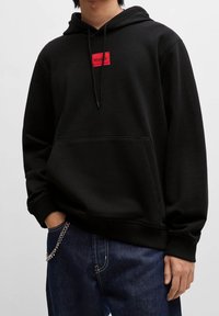 Persona con una sudadera negra con un parche rojo con el logotipo "HUGO", jeans azul oscuro y una cadena plateada que cuelga de un bolsillo.