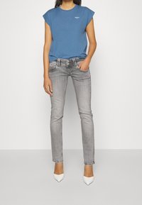 Camiseta corta de manga azul con un pequeño logo, combinada con jeans ajustados de color gris claro. El modelo lleva puestos unos tacones blancos de punta.